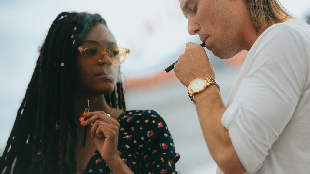 Two people smoking distillate vape pens outdoors, one wearing sunglasses and the other a gold watch
