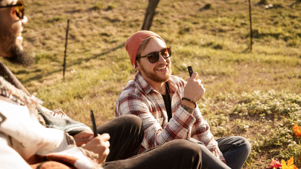 Two people relaxing outside on a sunny fall day, one holding a cannabis vape pen and smiling, enjoying autumn cannabis products.