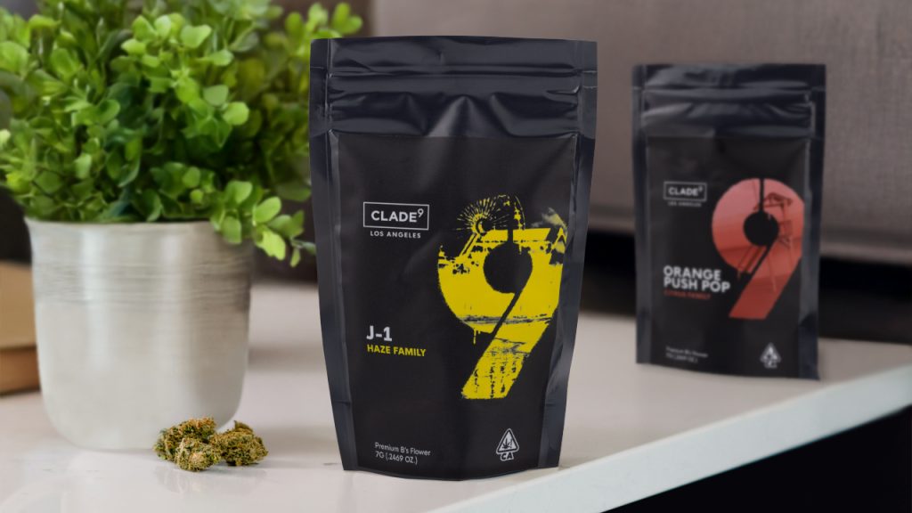 Clade9 cannabis flower packaging for J-1 and Orange Push Pop strains on a tabletop, with cannabis buds and a potted plant in the background.