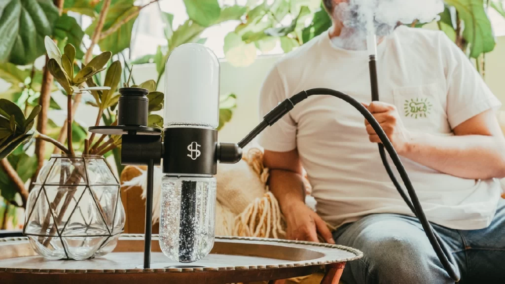 Modern glass gravity bong on a tray with a person exhaling vapor in the background.