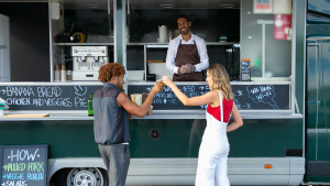 A smiling food truck vendor serves two happy customers outdoors at a local event in New Jersey